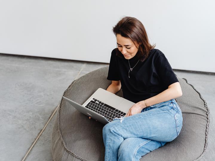 Smiling woman in black top sitting on a cushion, looking at her laptop.