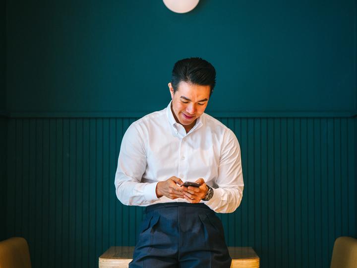 Man in white collared shirt leans against a table while looking happily at his cell phone.