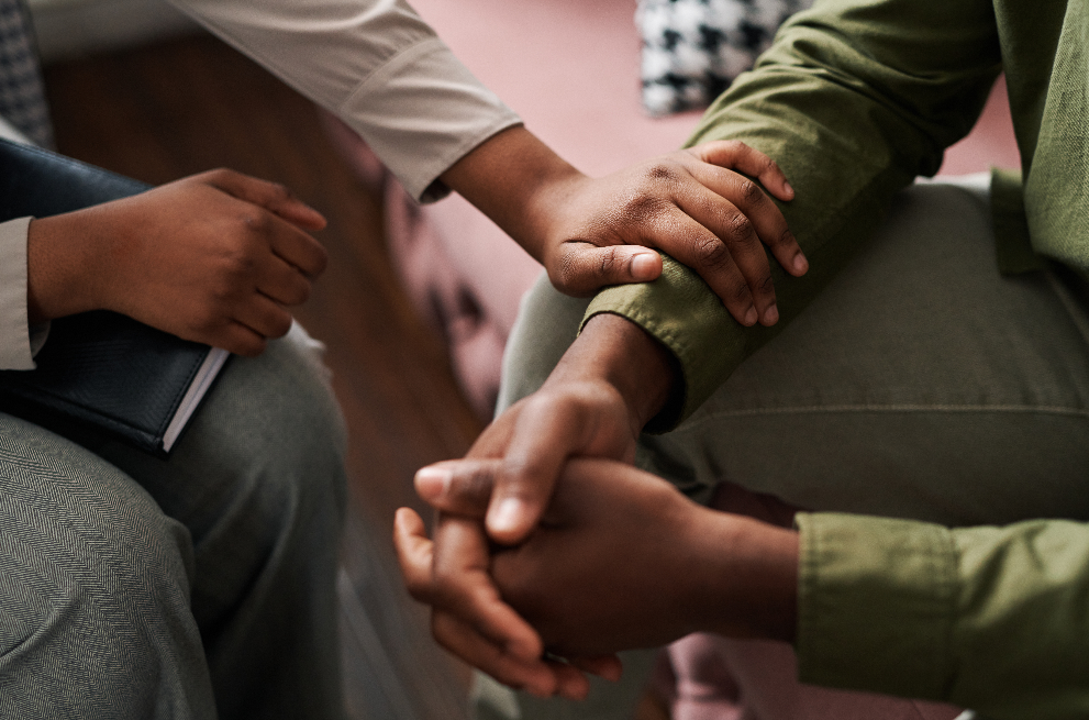  moment of quiet empathy and support. One person rests a hand gently on another’s arm, while the other sits with clasped hands