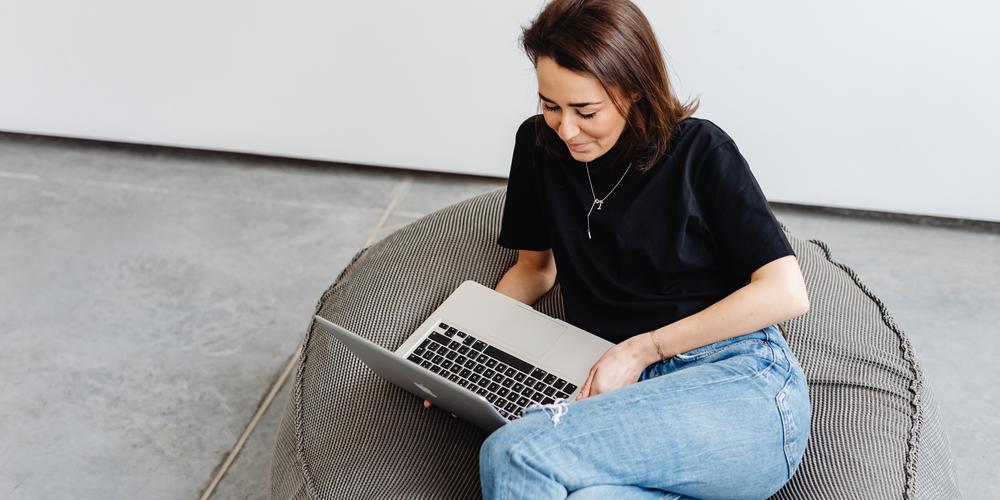 Smiling woman in black top sitting on a cushion, looking at her laptop.