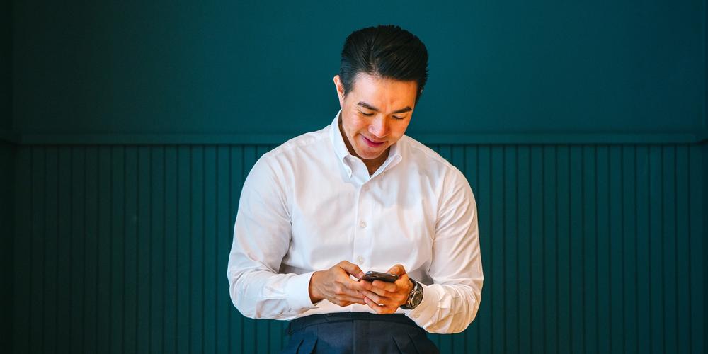 Man in white collared shirt leans against a table while looking happily at his cell phone.